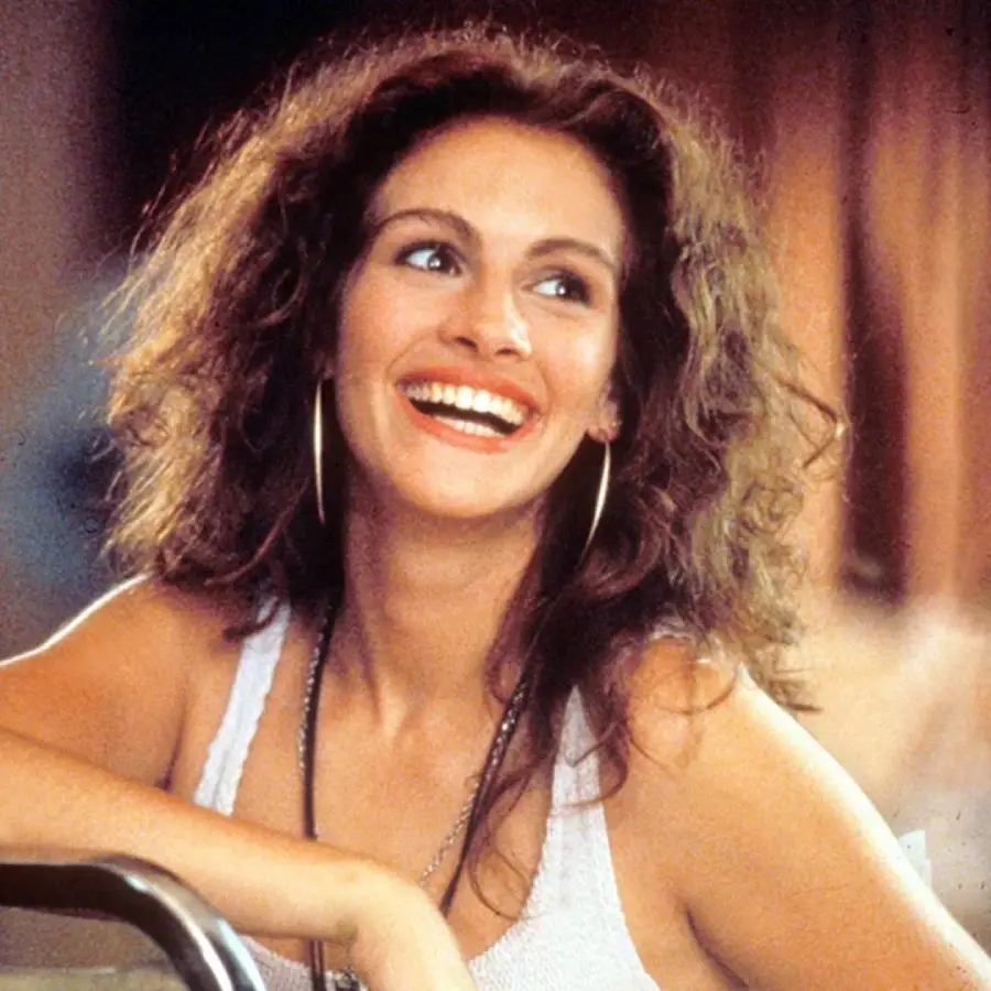 A woman with curly brown hair, wearing a white tank top and large hoop earrings, smiles brightly while looking slightly off to the side. The background is softly blurred.