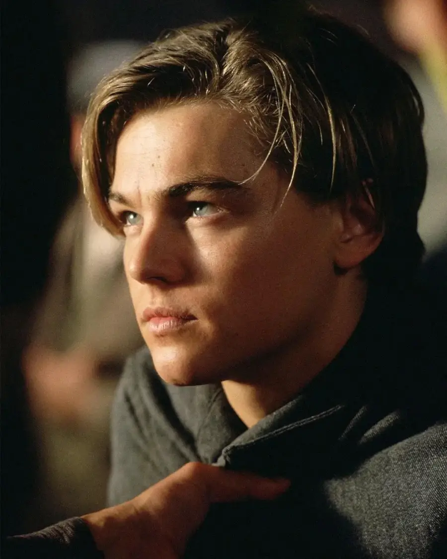 A young man with light brown hair and blue eyes gazes upward with a serious expression. He is wearing a gray shirt and is lit softly from one side, with a blurred background.
