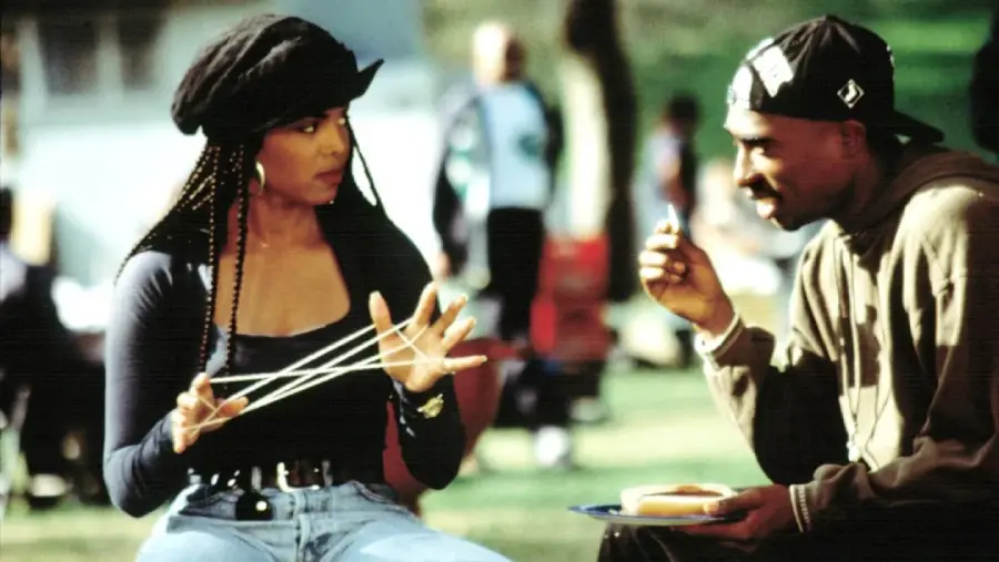 A woman with braids plays cat’s cradle with string while talking to a man in a cap who is holding a hot dog on a plate. They are sitting outdoors in a park, with people and greenery in the background.