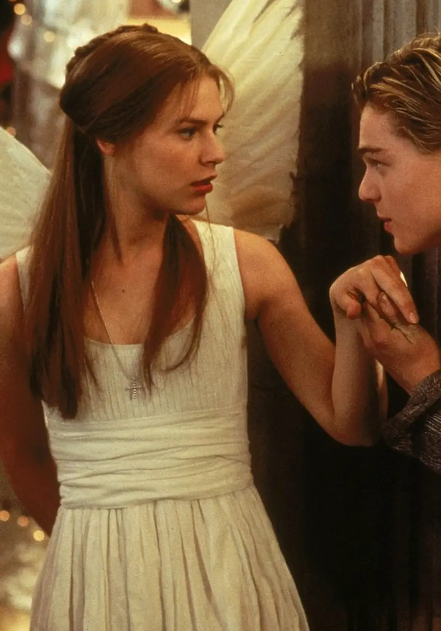 A young woman in a white dress and angel wings looks at a young man who is kissing her hand, in a dramatic scene from a film.