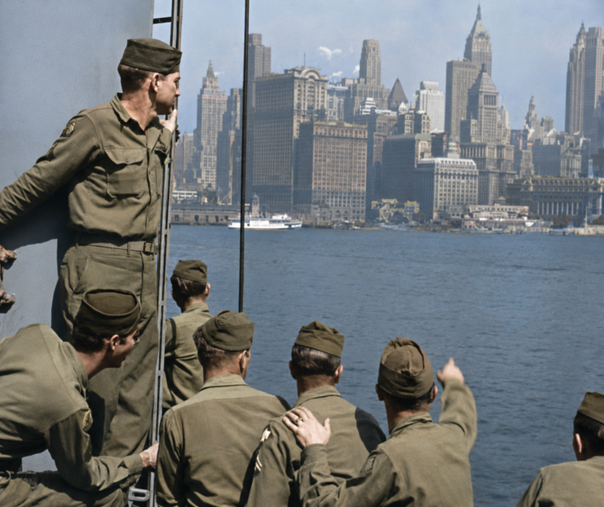 A group of soldiers in uniform stand on a ship, looking toward the skyline of New York City across the water on a clear day. One soldier points toward the city’s tall buildings.