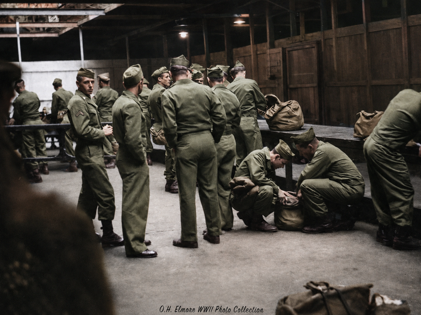 A group of WWII soldiers in green uniforms stand and kneel in a wooden barracks, sorting through duffel bags on long tables and the floor. The scene is busy, with soldiers preparing or packing belongings.