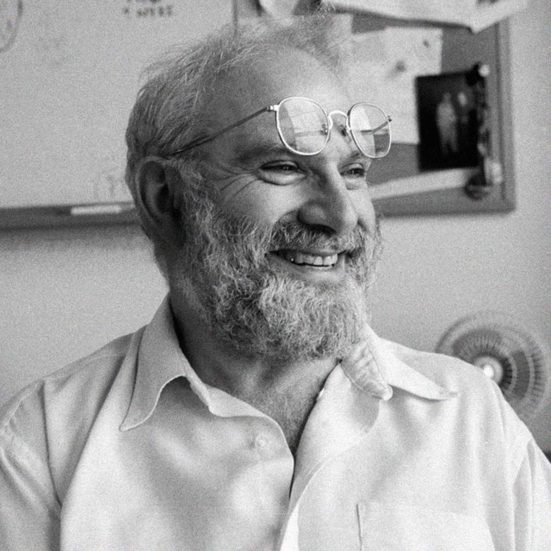 A bearded man with glasses perched on his forehead smiles while sitting in front of a bulletin board covered with papers and photos in a black-and-white photo.