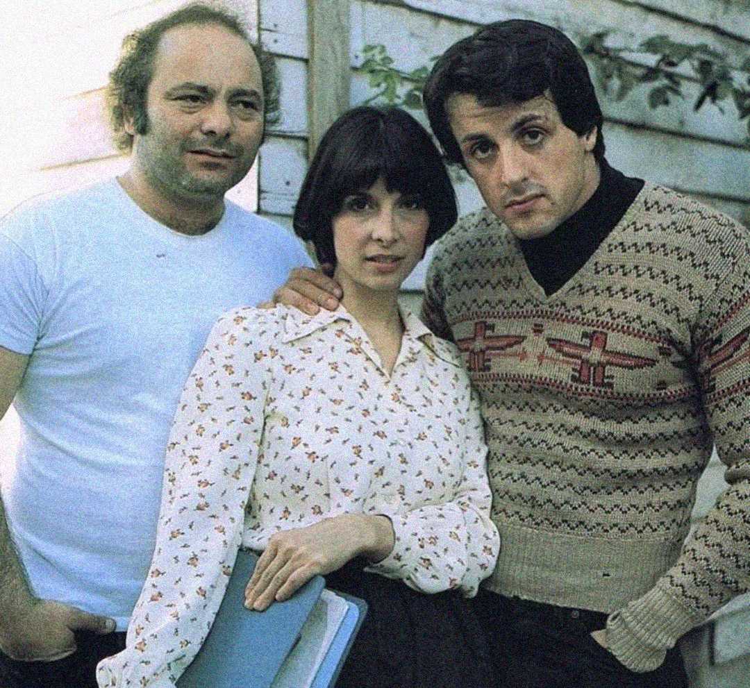 Three people pose together outside near a wooden wall. The man on the left wears a white t-shirt, the woman in the middle holds a blue folder and wears a patterned blouse, and the man on the right wears a patterned sweater.