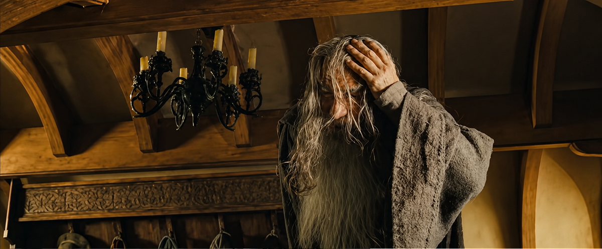 An elderly man with long gray hair and a beard stands indoors, wearing a robe and holding his head with one hand as if in distress or deep thought. A chandelier and wooden beams are visible in the background.