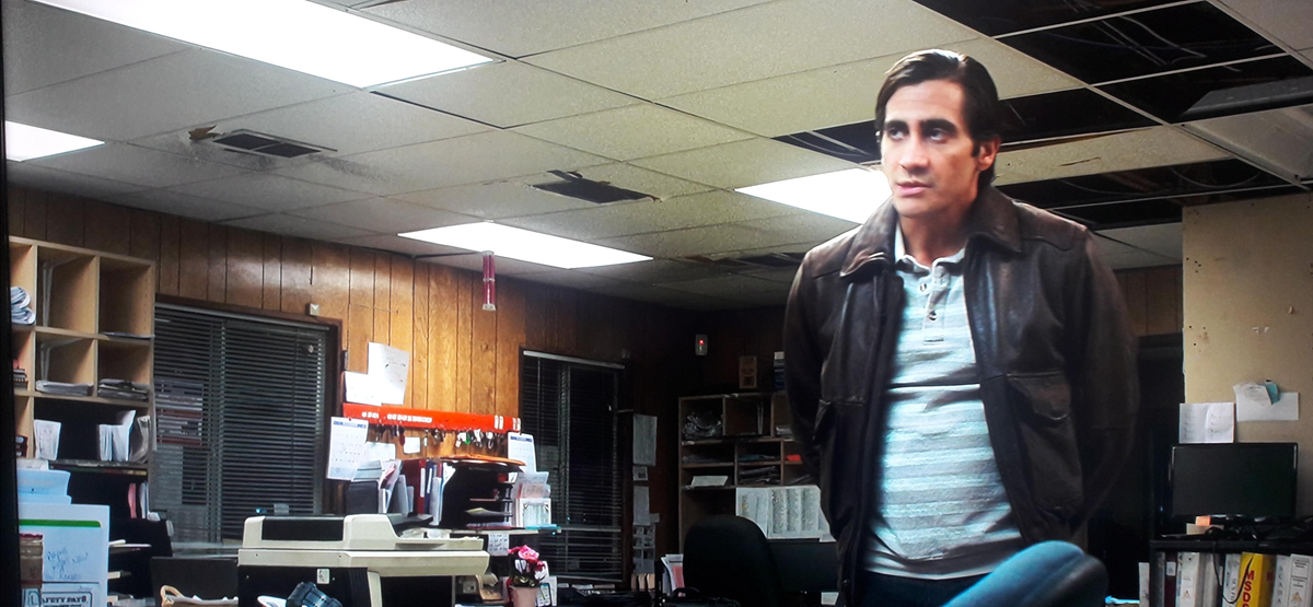 A man in a leather jacket stands in a cluttered office with wood-paneled walls, messy desks, paperwork, and ceiling tiles missing or damaged.