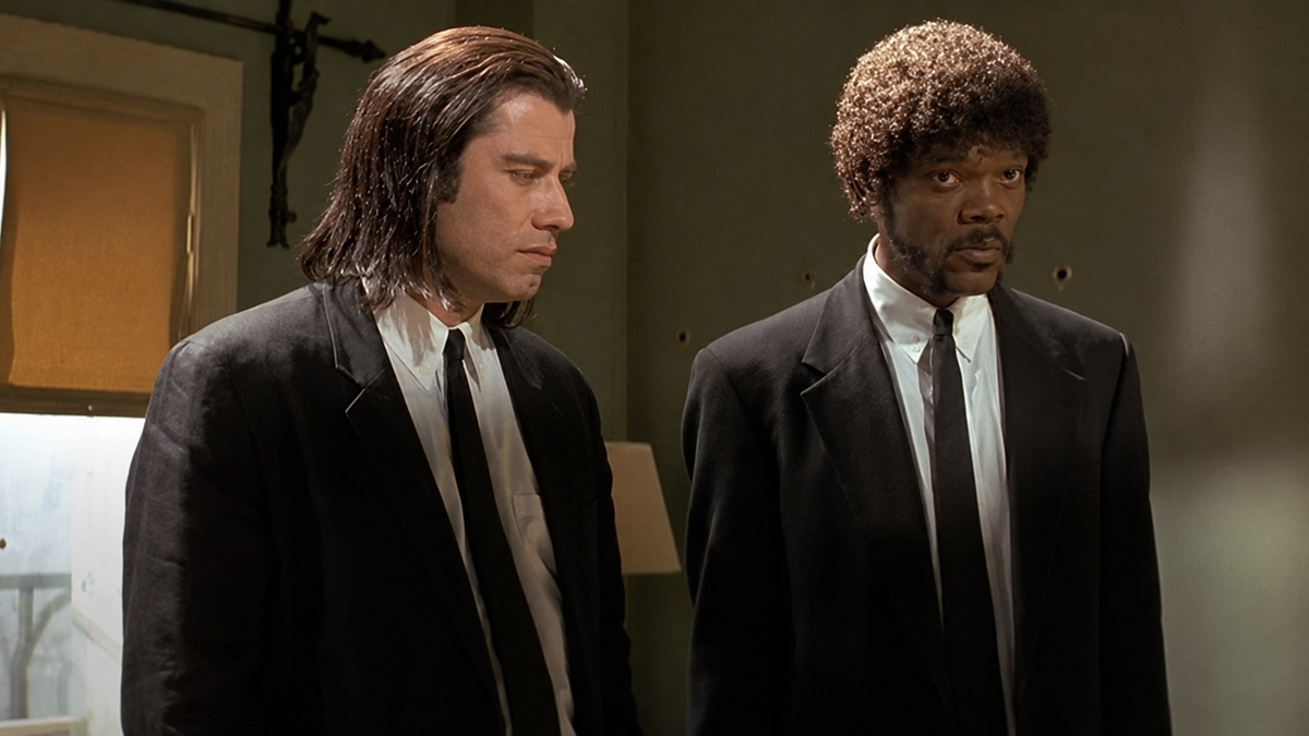 Two men in black suits and white shirts stand side by side in a dimly lit room. One has long straight hair, the other has curly hair and a goatee. Both have serious expressions and are looking slightly downward.