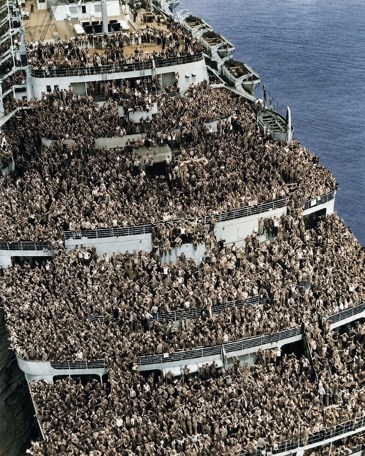 A large ship at sea is densely packed with thousands of uniformed soldiers on multiple decks, all gathered closely together, many waving towards the camera. The ocean is visible in the background.