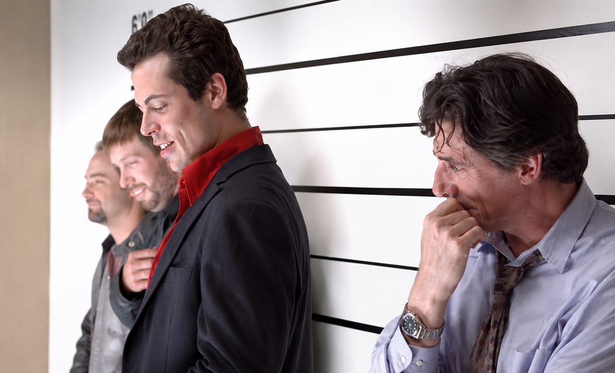 Four men stand in front of a police lineup height chart, laughing and smiling, with three looking left and one looking down while covering his mouth with his hand. All are casually dressed.
