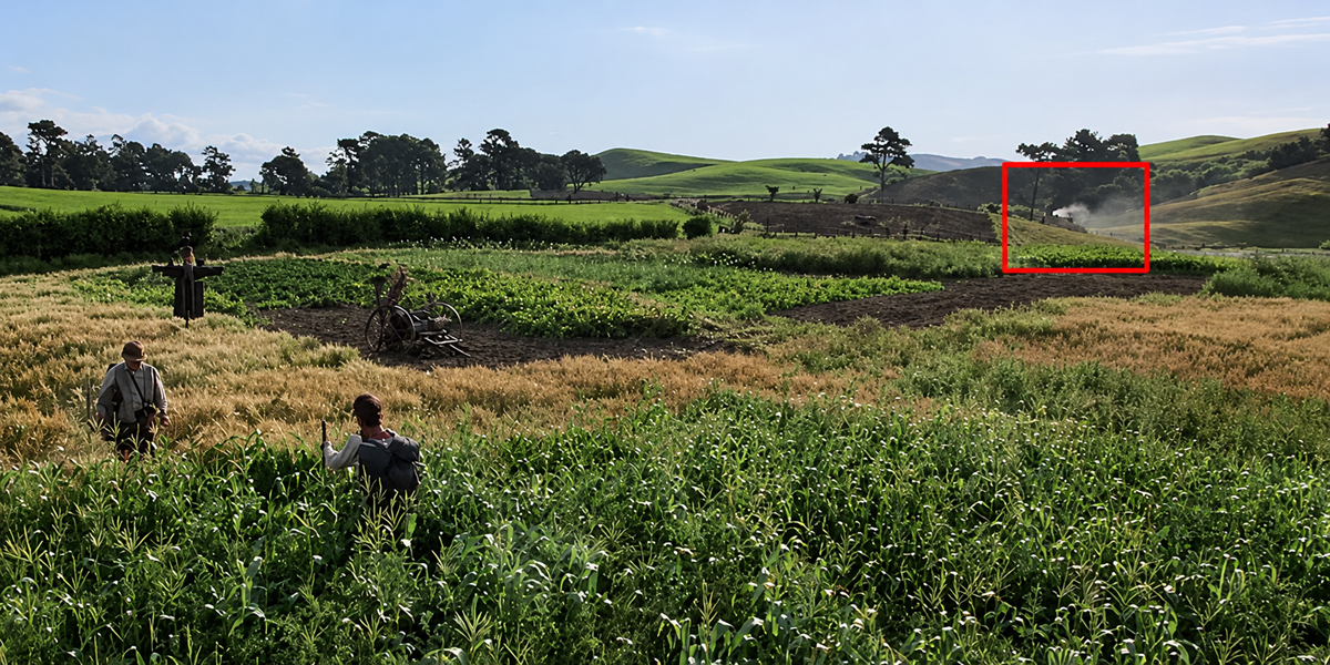 Two people walk through lush green fields with crops, set in a rural landscape. There are hills, trees, and two small houses partially visible in the background. The sky is clear and blue.