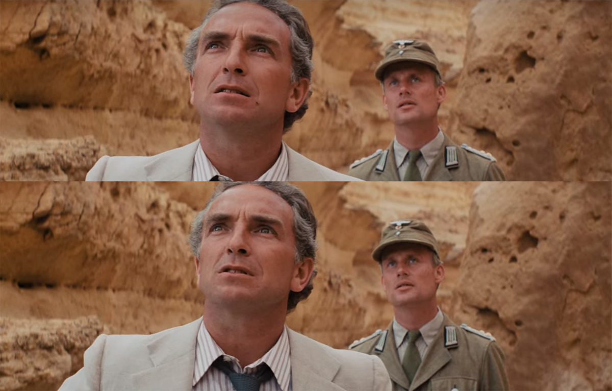 Two men stand outdoors in front of rocky cliffs. The man in front wears a light suit and tie, looking up with concern. Behind him, a man in a tan military uniform and cap also looks upward, with an uncertain expression.