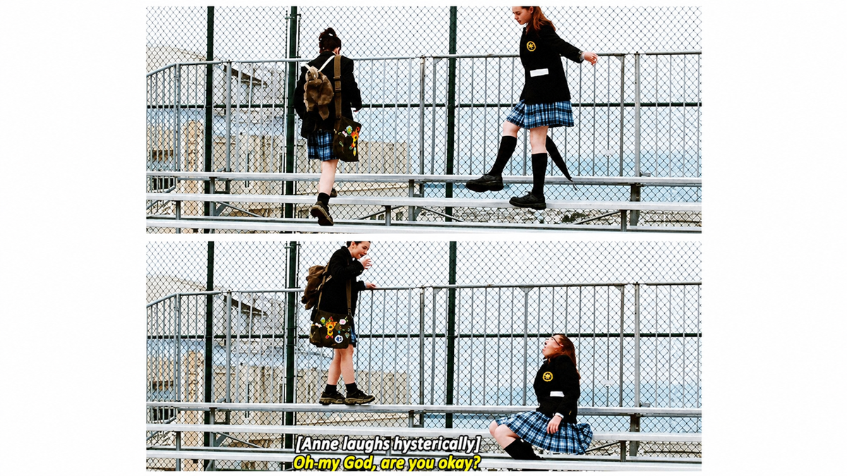 Two girls in school uniforms interact on metal bleachers. One girl sits on a higher row laughing, while the other stands looking at her. The subtitle reads, "Oh my God, are you okay?".