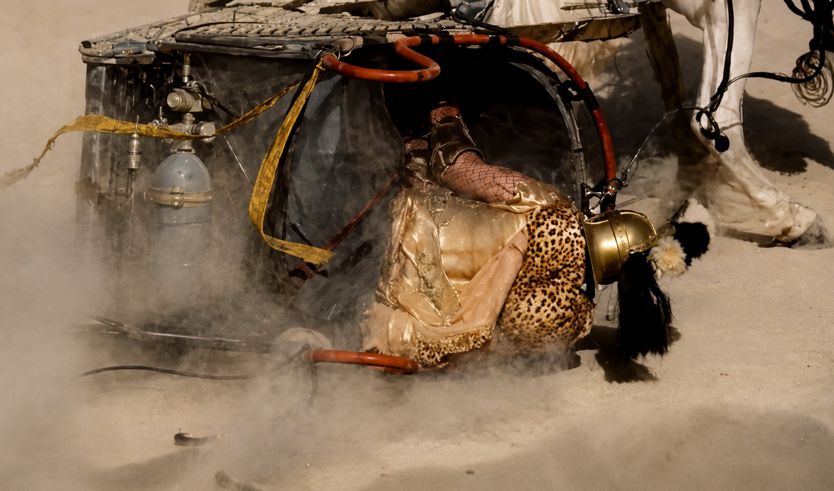 A person in a gold helmet and leopard print outfit is partially under a toppled chariot on a sandy surface, surrounded by dust and harnesses, with a white horse visible in the background.