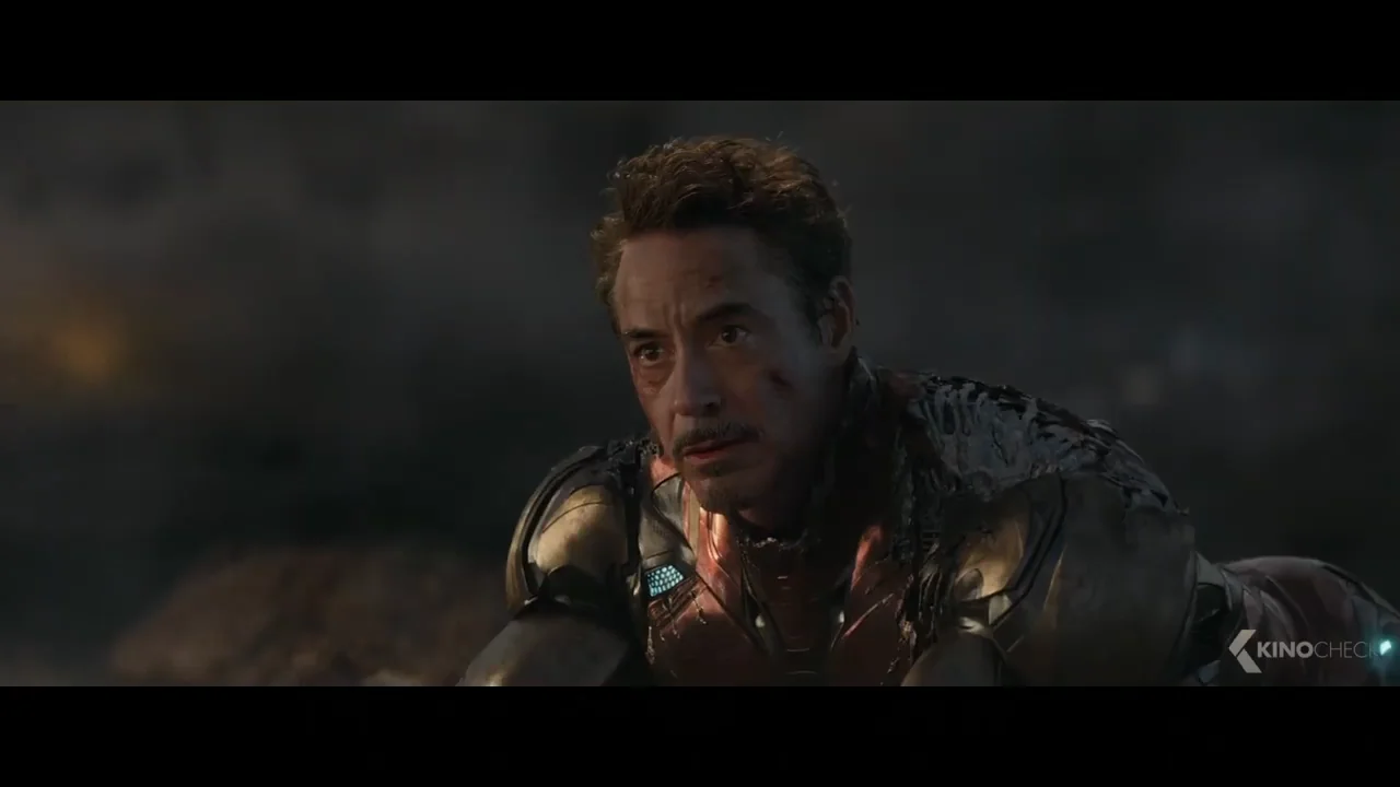 A man in a damaged metallic suit kneels on the ground, looking up with a serious, exhausted expression. His face and armor are dirty and battered, and the background is dark and smoky.