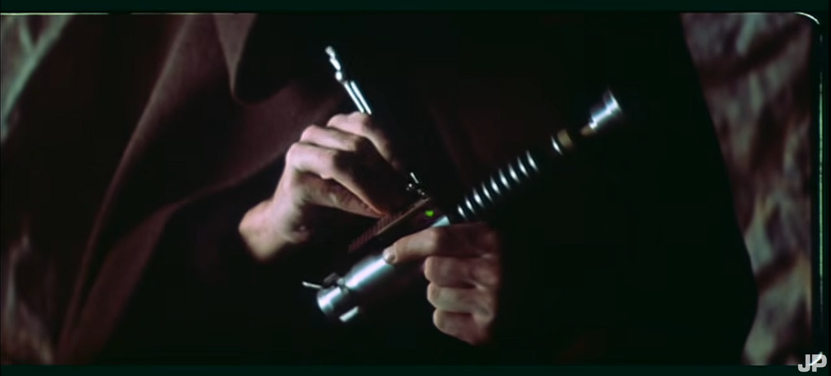 A person in a dark cloak adjusts a metallic lightsaber hilt, holding it with both hands against a dim background.