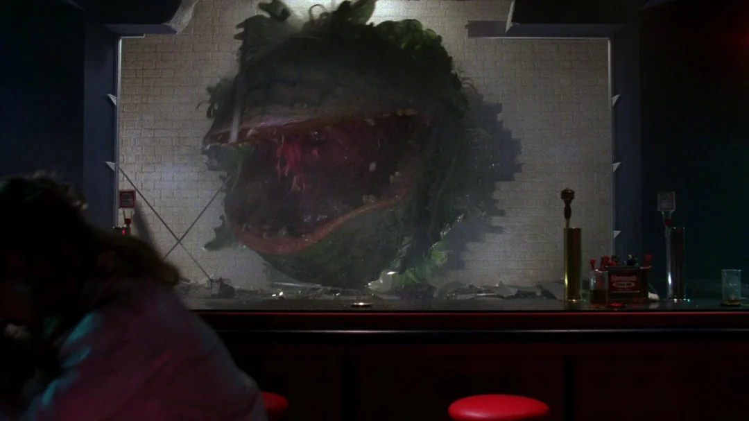 A giant, menacing plant with sharp teeth bursts through a tiled wall behind a bar counter, while a person appears blurred in the foreground and two red barstools sit in front of the counter.