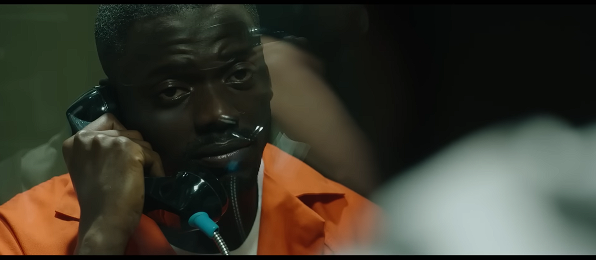 A man in an orange prison uniform holds a phone to his ear, sitting behind glass, appearing somber during a visitation in a prison setting.