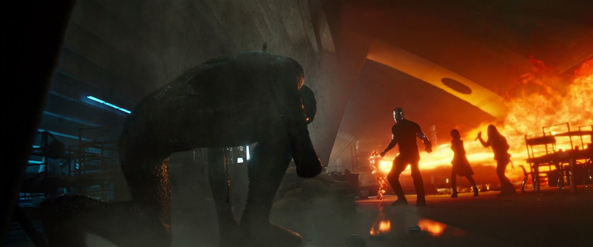 A large, shadowy creature crouches in the foreground of an industrial, dimly lit space. In the background, three people stand near bright flames and smoke, suggesting chaos or a confrontation.