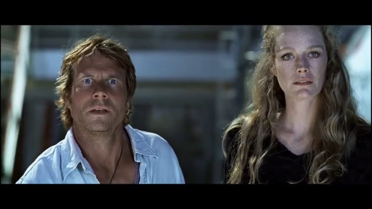 A man and a woman stand side by side indoors, both looking ahead with serious and intense expressions. The background is blurred, focusing attention on their faces. Movie.