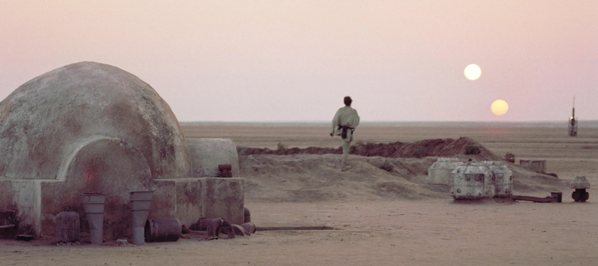 A person stands on a sandy desert near a round, dome-like structure at sunset, looking at two suns in the sky. The landscape is barren and flat, with scattered objects and soft, pinkish light.