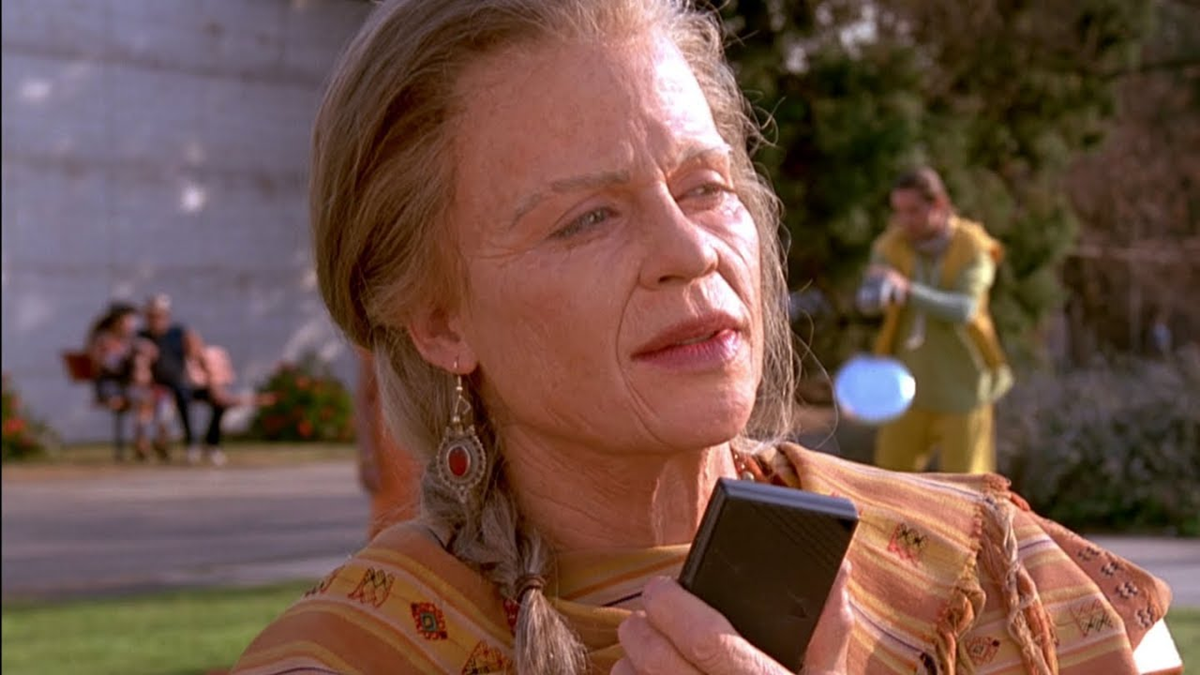 An older woman with braided hair and patterned clothing holds a small black device, standing outdoors. A person in yellow is blurred in the background near trees and benches.