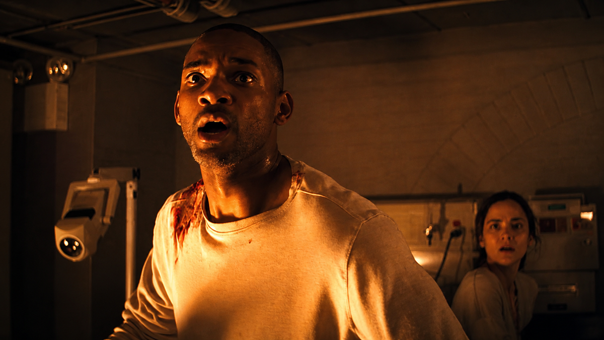 A man with a wounded shoulder and a shocked expression stands in a dimly lit room, while a concerned woman sits behind him near medical equipment.