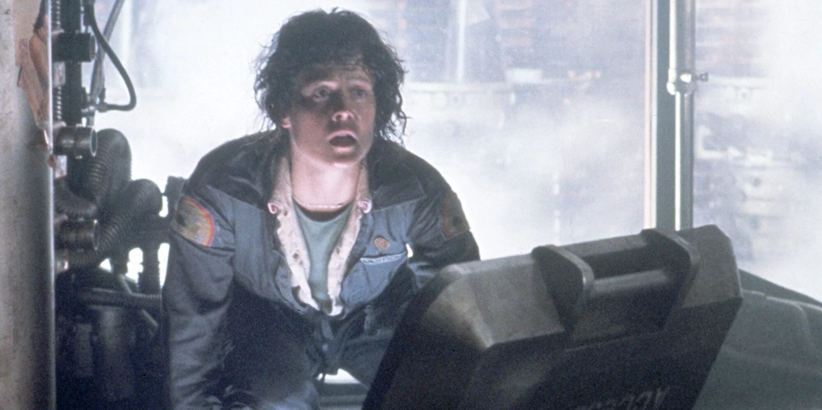 A person with short, messy hair and a distressed expression wears a blue jumpsuit, kneeling in a smoky, industrial setting, illuminated by harsh light and looking upward in alarm.