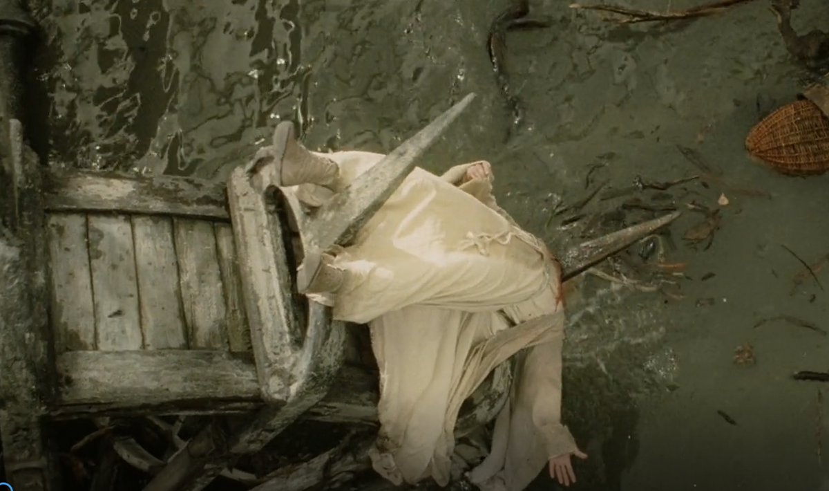 A person in light-colored clothing lies unconscious on a broken wooden structure in muddy water, surrounded by debris and a wicker basket.