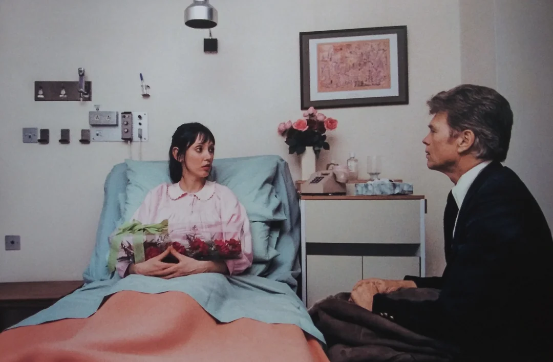 A woman in a hospital bed wearing a pink gown holds flowers and looks at a man sitting beside her. There are medical devices on the wall, a vase of roses, and a framed picture above the bed.