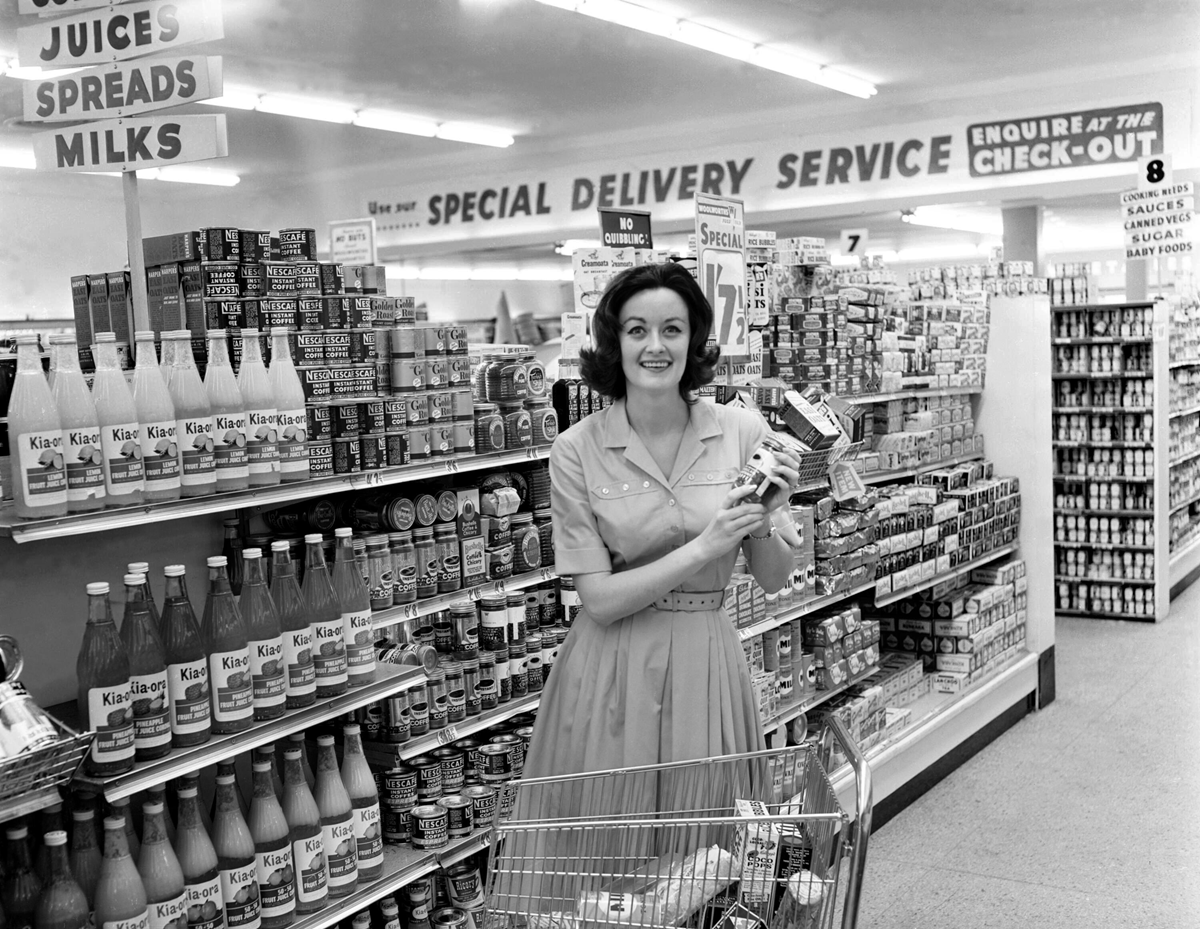 A woman in a vintage dress stands in a grocery store aisle, smiling and holding boxed groceries. Shelves are stocked with bottles, cans, and jars. A sign above reads "JUICES SPREADS MILKS" and another says "SPECIAL DELIVERY SERVICE.