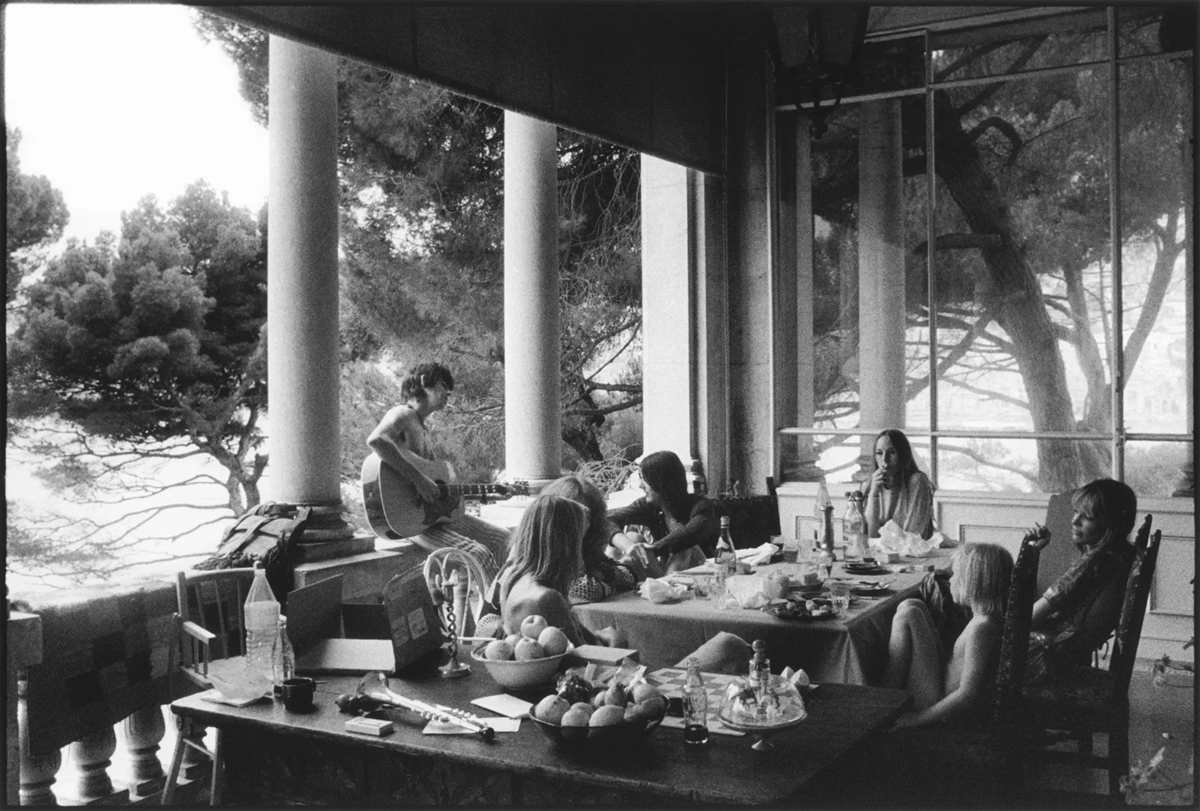 A group of people relax on a porch, some sitting around a table with food and drinks, while one person plays a guitar near large columns and trees in the background. The scene is in black and white. 1. The Rolling Stones, Exile on Main St. Album