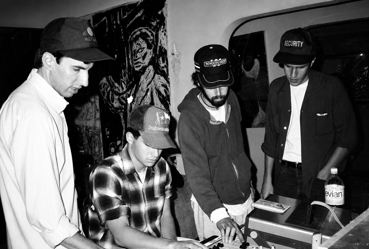 Four young men wearing baseball caps gather around electronic equipment in a dimly lit room. One is operating a keyboard or controller, while the others look on. A water bottle and wall art are visible in the background. 8. Beastie Boys, Paul’s Boutique