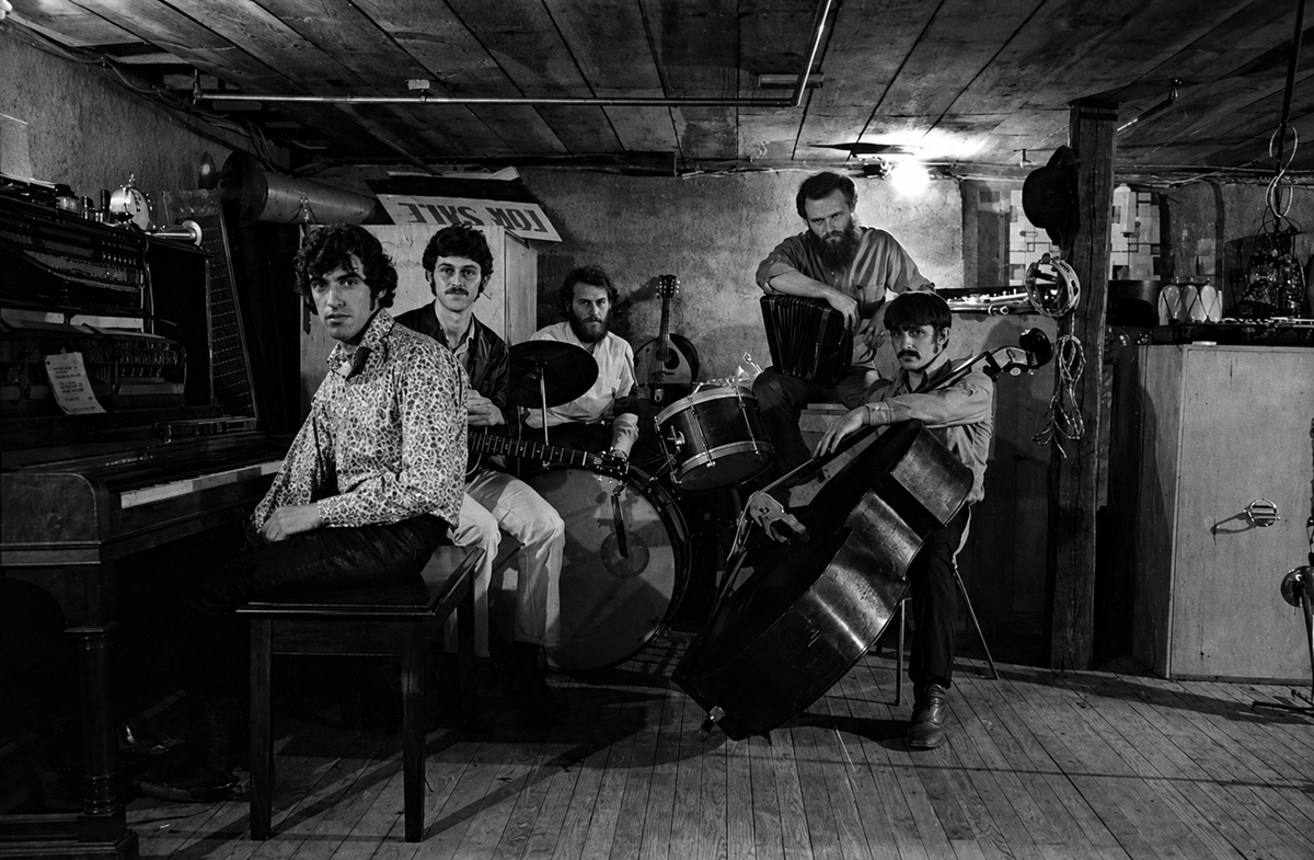 Five men with musical instruments, including piano, guitar, drums, accordion, and upright bass, pose in a dimly lit, rustic basement with wooden floors and ceiling. The mood is serious and artistic. 6. Bob Dylan and The Band, The Basement Tapes