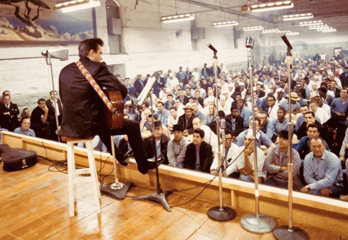 A musician with a guitar performs on stage for a large seated audience in blue uniforms inside a prison-like hall, with multiple microphones set up in front of him. 4. Johnny Cash, At Folsom Prison