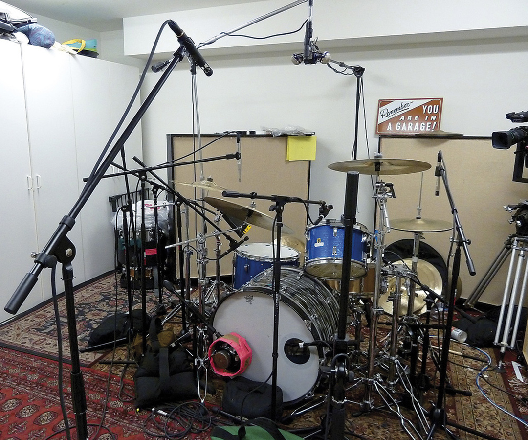 A blue drum set is surrounded by multiple microphones on stands in a home studio with white cabinets, soundproof panels, and a patterned rug. A sign on the wall reads, “Remember, you are in a garage!” 10. Foo Fighters, Wasting Light