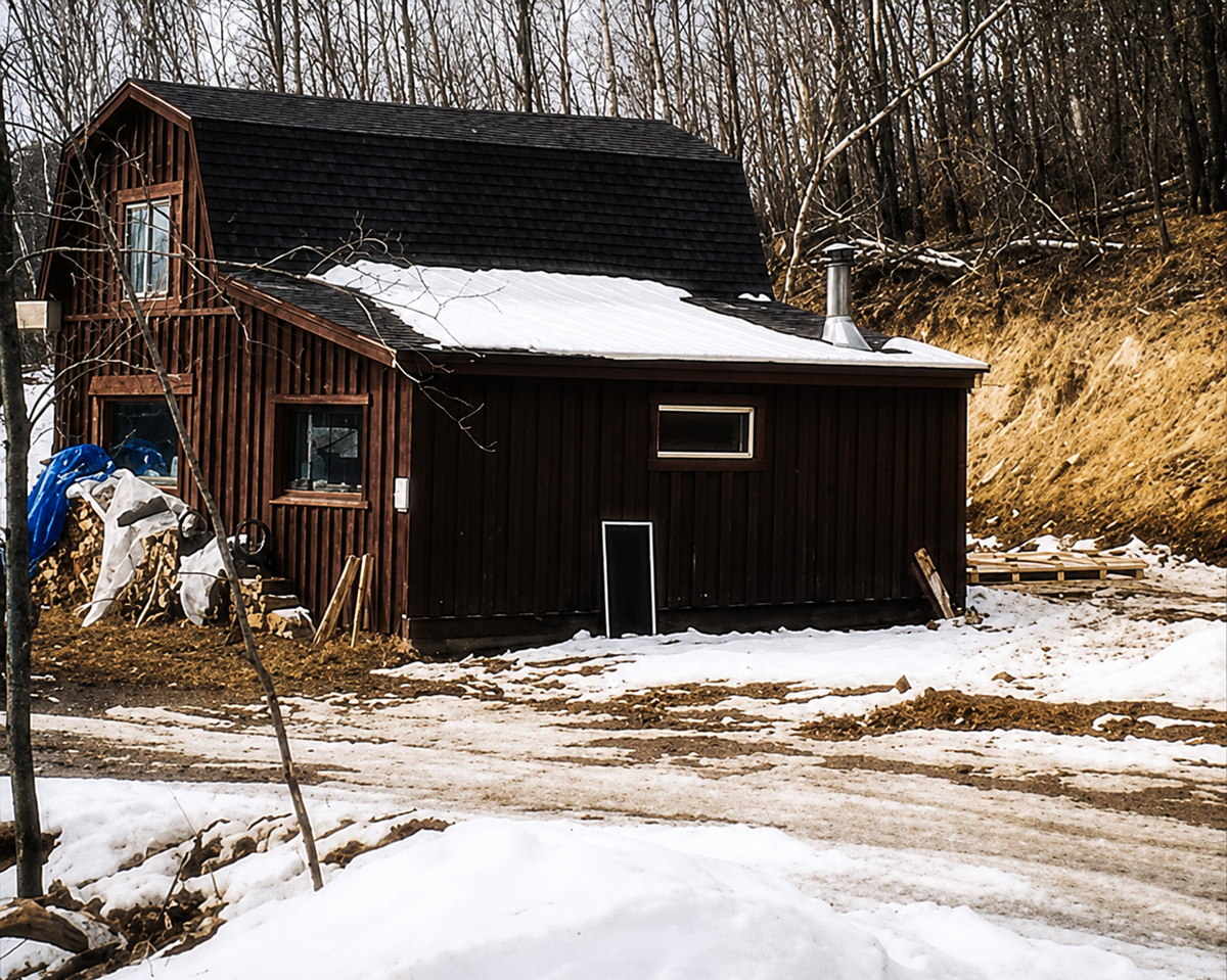 A dark brown wooden house with a sloped roof sits on a snowy landscape. Bare trees surround the house, and construction materials are scattered nearby. The ground is partially covered with snow and patches of dirt. 2. Bon Iver, For Emma, Forever Ago