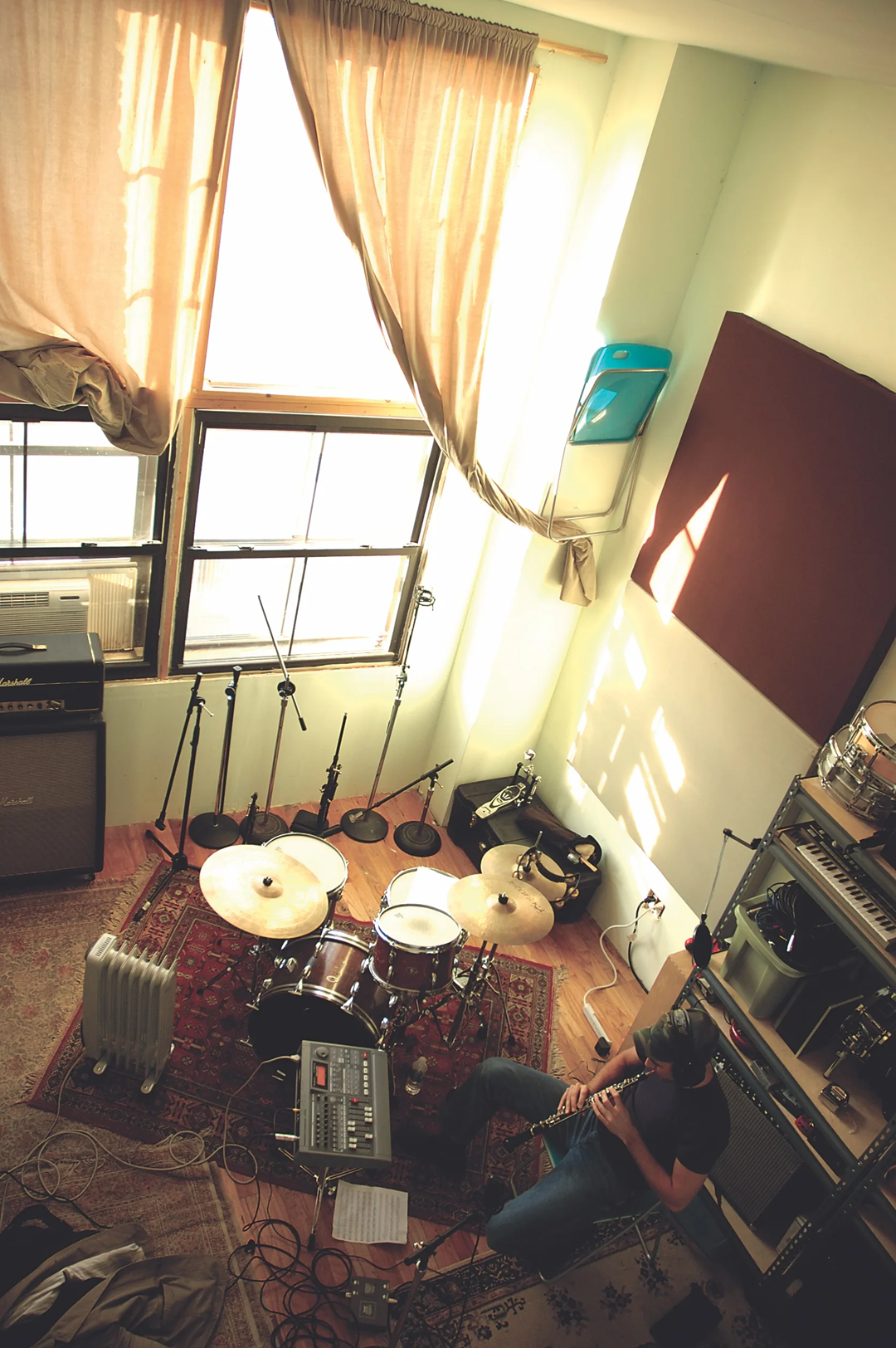 A person sits playing guitar in a sunlit music studio with a drum set, amplifiers, a radiator, and various microphones on stands. Light streams through large windows with beige curtains. 12. Sufjan Stevens, Michigan