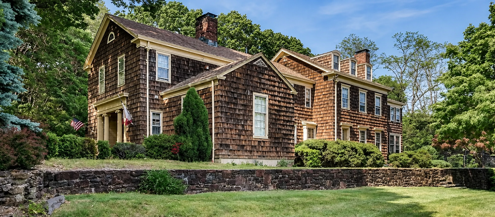 A large, historic house with brown wooden shingles, white-trimmed windows, and multiple chimneys, surrounded by green trees, bushes, and a stone retaining wall under a blue sky. 3. Bruce Springsteen, Nebraska