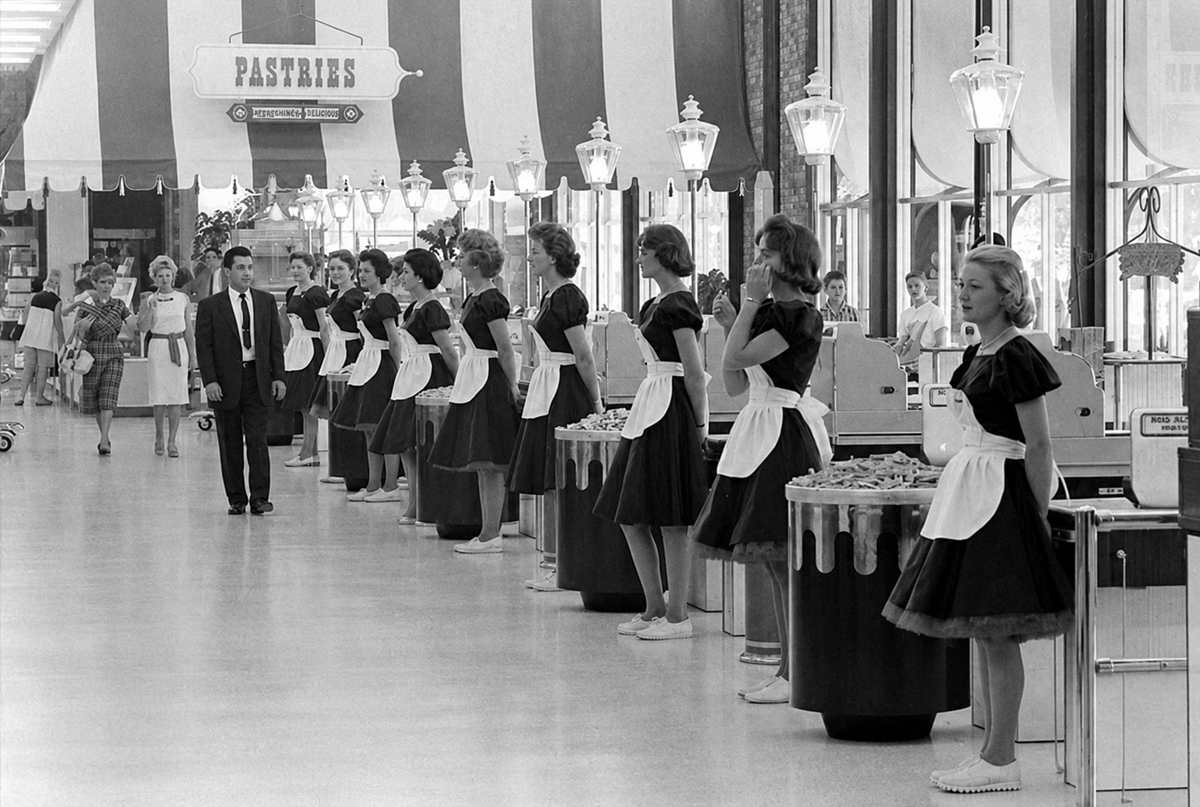 A row of women in matching dresses and aprons stand behind counters in a vintage store, with pastries sign above. A man in a suit walks past, and shoppers are visible in the background.