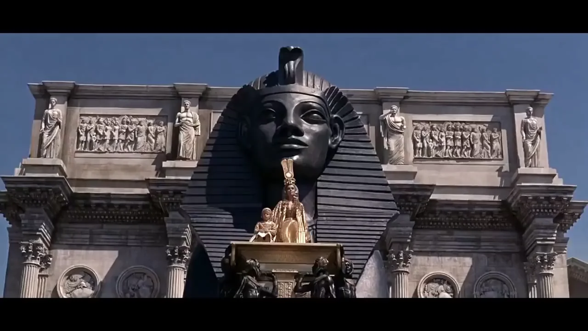 A large, black Egyptian pharaoh statue is flanked by carvings and topped by a gold statue of seated figures against a clear blue sky, resembling ancient Egyptian architecture. 4. Cleopatra’s Giant Roman Entrance