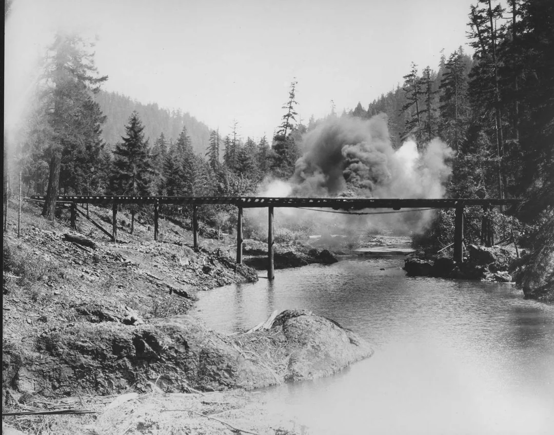 A wooden bridge over a river in a forested area is partially obscured by a large cloud of smoke, likely from an explosion, with trees and hills in the background. 7. The General Collapsing Bridge