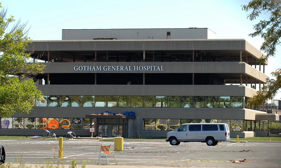 A partially destroyed building labeled "Gotham General Hospital" with broken windows and debris outside; a white van is parked in front on an empty lot. 3. The Dark Knight’s Gotham General Hospital