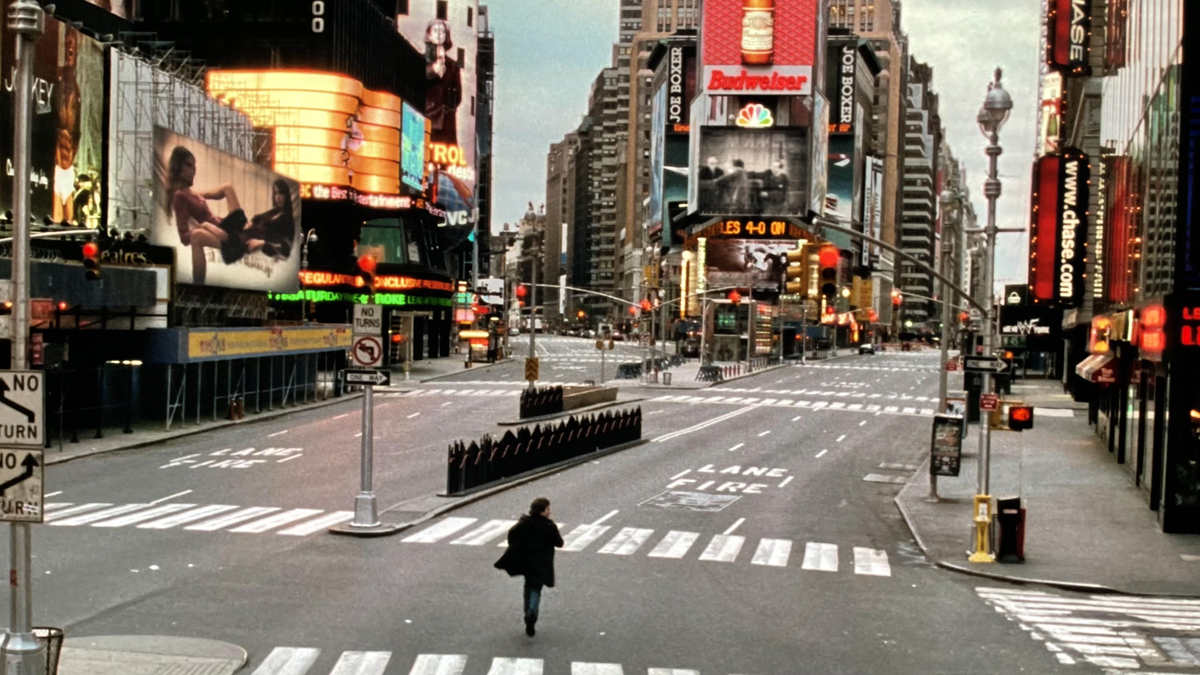 A person walks alone across an empty city street surrounded by tall buildings, bright billboards, and traffic lights in what appears to be Times Square, New York City. The scene is quiet and deserted. 8. Vanilla Sky Empty Times Square