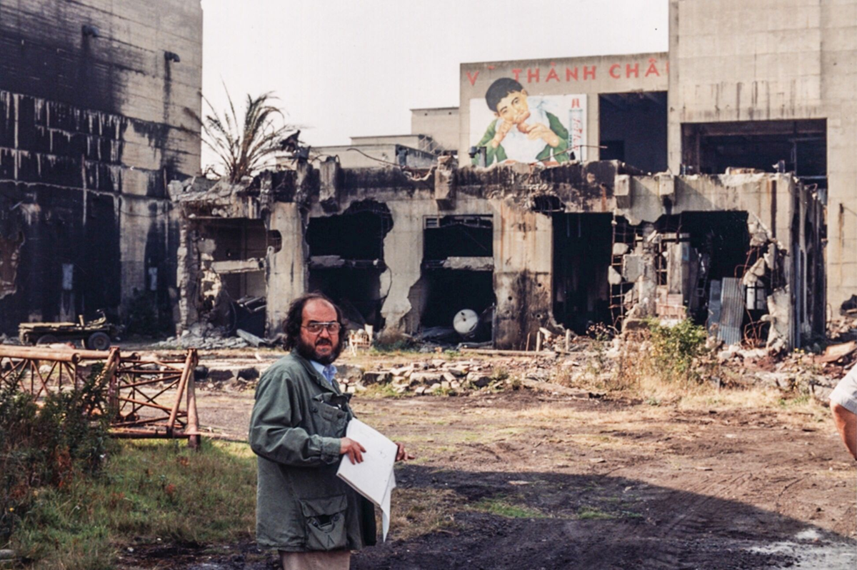 A man holding papers stands in front of a heavily damaged, burned building with a mural of a child eating on an adjacent wall. Debris and overgrown plants cover the ground around the ruins. 9. Full Metal Jacket Bombed-Out Huế