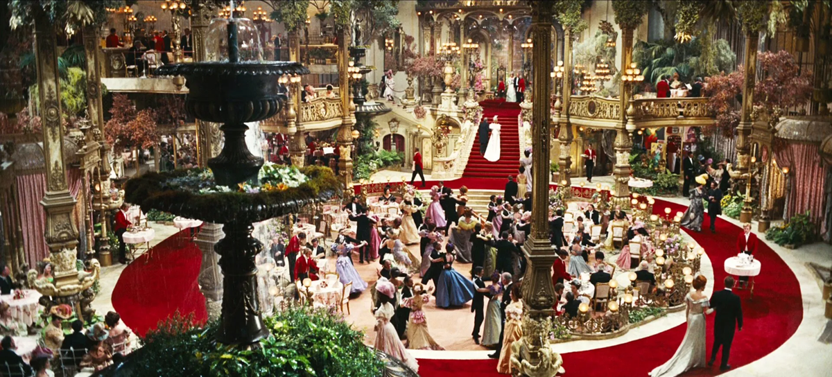 A lavish ballroom scene with elegant guests in formal attire, ornate golden decor, a grand staircase, a large fountain, and tables set for a celebration on a red carpet. 10. Hello, Dolly! Harmonia Gardens Restaurant