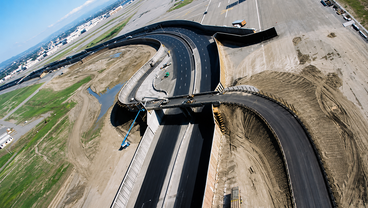 Aerial view of a highway overpass under construction, with newly paved roads and construction vehicles present. Surrounding areas show dirt, grass, and equipment, indicating ongoing infrastructure development. 1. The Matrix Reloaded Private Freeway