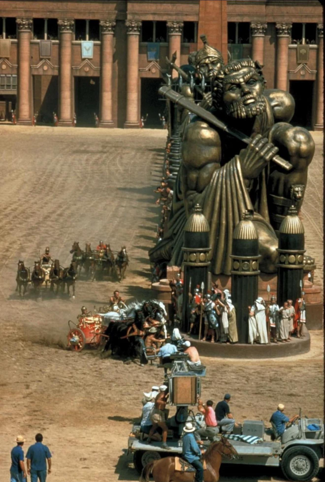 A scene set in ancient Rome features large statues of muscular men with swords, chariots racing on dirt, actors in period costumes, and a film crew directing from modern equipment in the foreground. 2. Ben-Hur’s Massive Chariot Arena