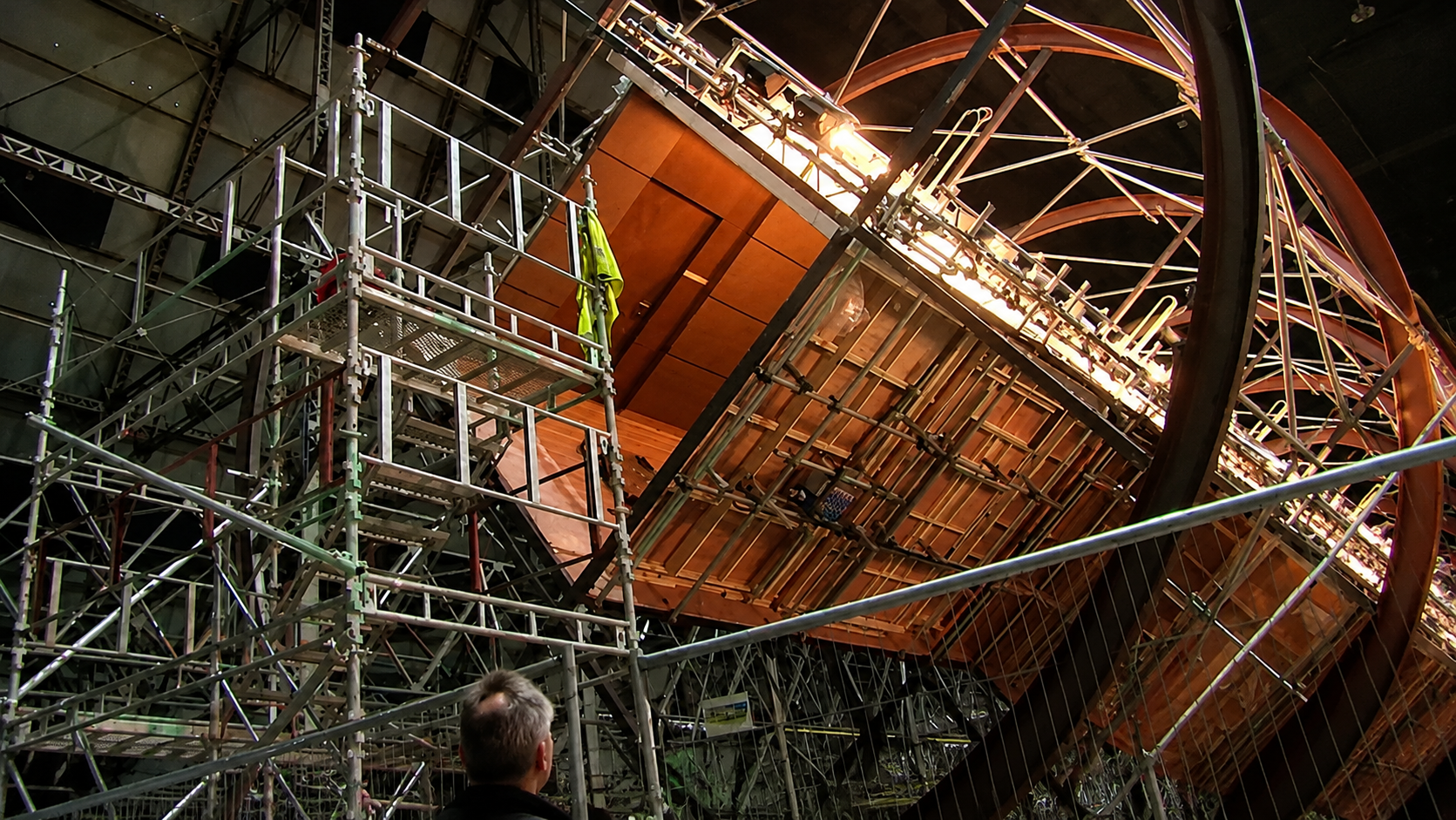 A person stands in front of a large, partially constructed rotating set surrounded by metal scaffolding, with bright lights illuminating the structure inside a spacious industrial building. 6. Inception Rotating Hotel Hallway