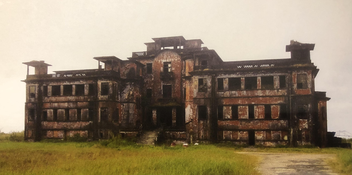 An abandoned, weathered brick building with broken windows and crumbling walls sits surrounded by overgrown grass under a cloudy sky. Stairs lead to a dark central entrance. 15. Le Bokor Palace, Bokor Mountain, Cambodia