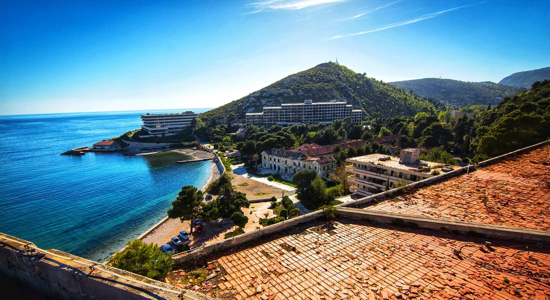 A coastal view with turquoise water, a sandy beach, green hills, and modern buildings surrounded by trees. In the foreground, there are old, weathered rooftops. The sky is clear and blue. 4. The Kupari Resort Complex, Croatia